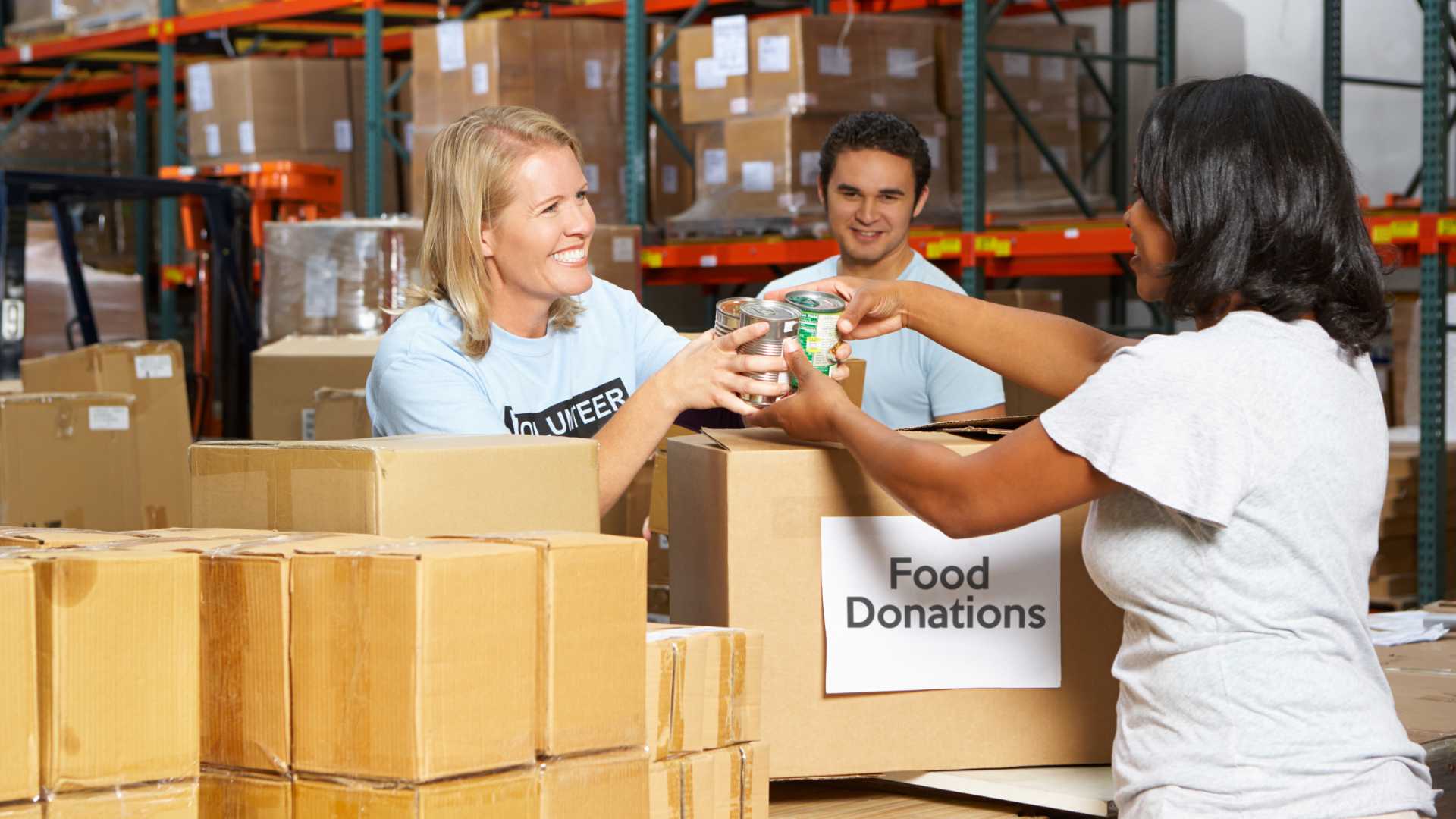 Two people wearing volunteer shirts standing in a warehouse with boxes around them labeled "food donations" handing out food to a person in need.