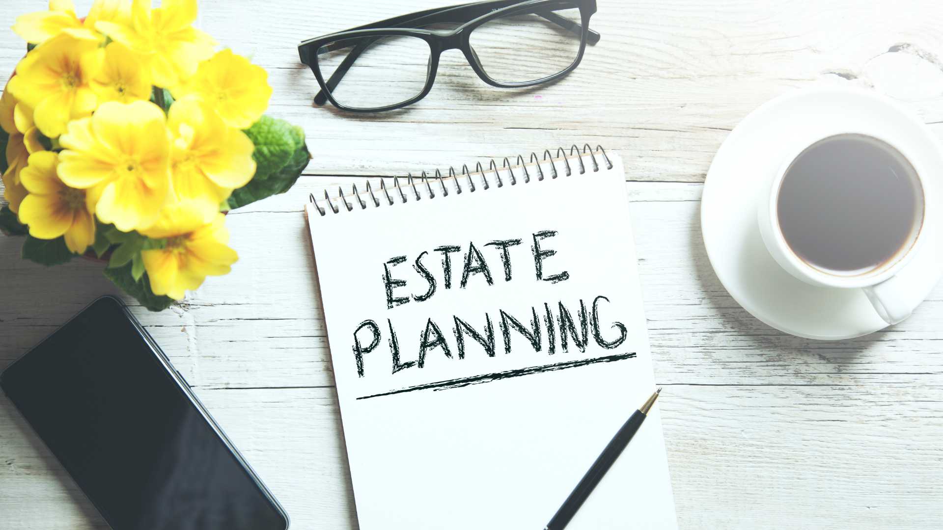 A white table with a vase of yellow flowers, a coffee cup, a pair of glasses, and a notepad and pen on it. The pen is lying on the notepad and the notepad says "ESTATE PLANNING" at the top.